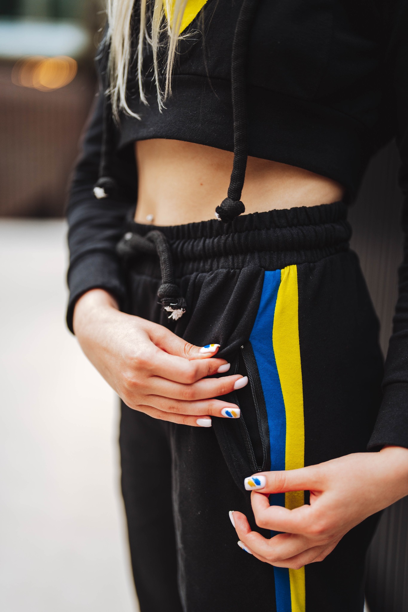 Fragment damskiego kompletu dresowego: czarne spodnie typu joggers z niebiesko-żółtym lampasem i rozpiętym zamkiem kieszeni, oraz krótka bluza z kapturem, eksponujące brzuch z piercingiem, dłonie...