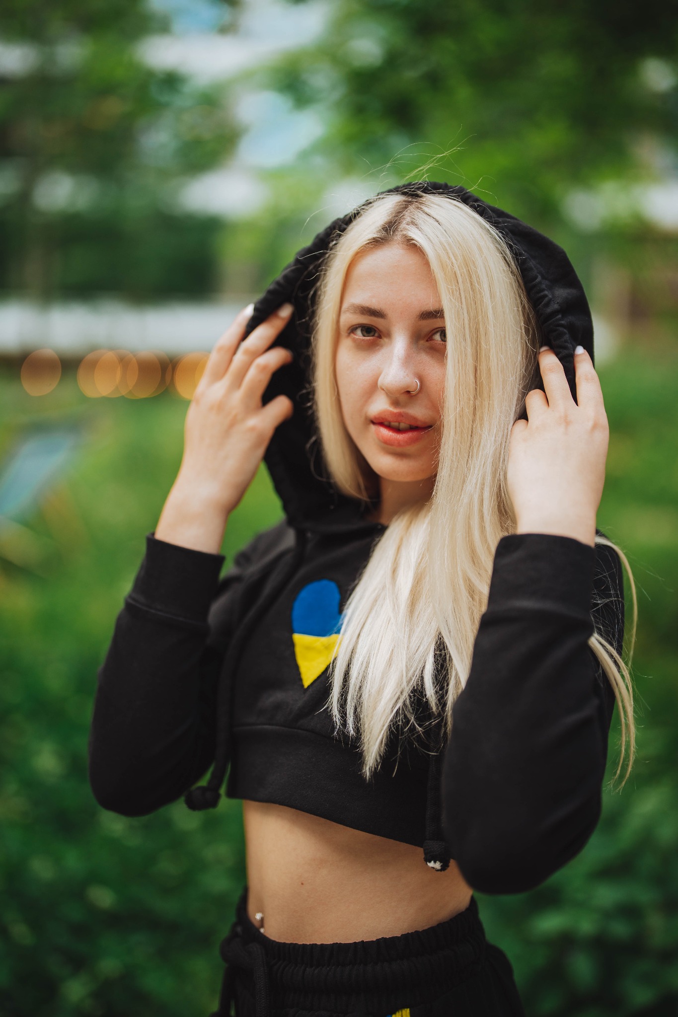 Portret młodej kobiety z blond włosami w czarnej bluzie z kapturem, ozdobionej niebiesko-żółtym symbolem, na tle zieleni.