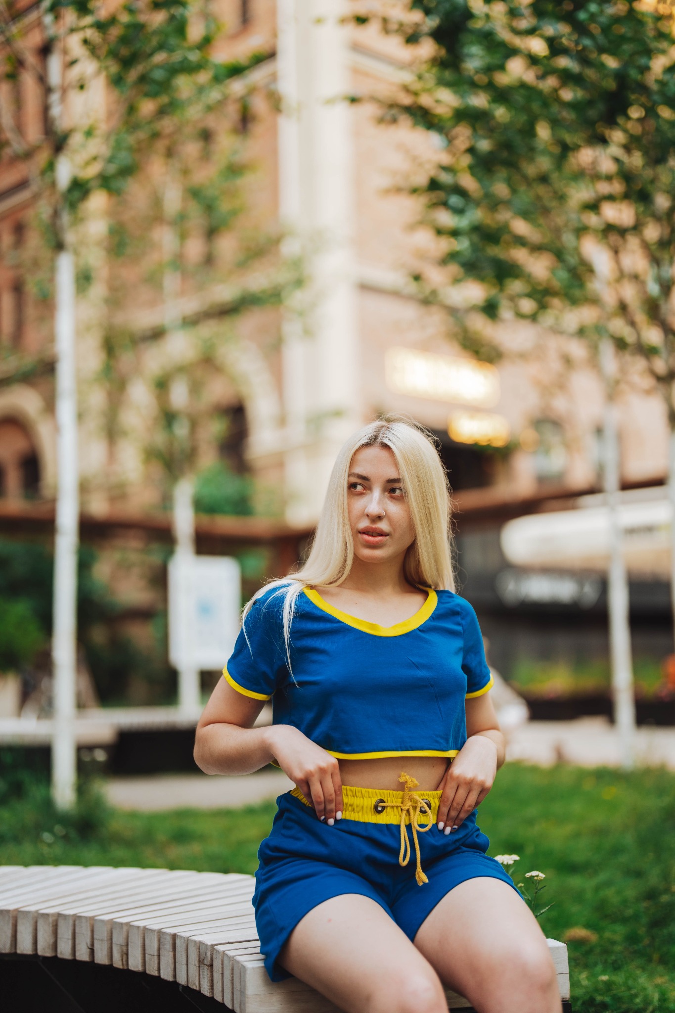 Młoda kobieta z blond włosami siedzi na ławce w parku, ubrana w niebieski komplet sportowy z żółtymi akcentami, spodenki z sznurkiem w pasie, patrzy w bok.
