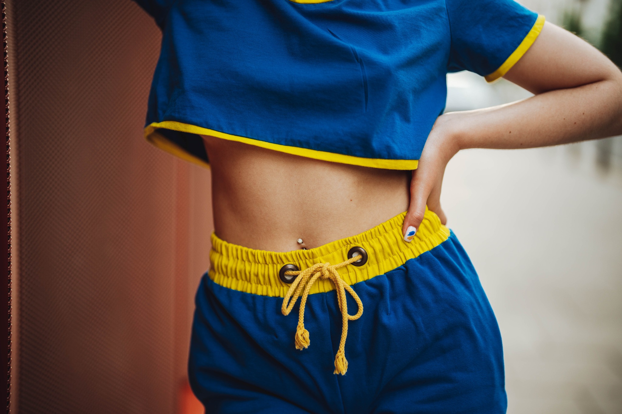 Krótki, niebieski top i szorty z żółtymi lamówkami, widoczny fragment torsu z piercingiem pępka, dłoń z paznokciem ozdobionym niebieskim wzorem.
