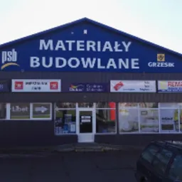 Fasada budynku z napisem 'MATERIAŁY BUDOWLANE' i logotypami różnych producentów, widoczne zaparkowane samochody i fragment zieleni w tle.