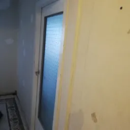 Ściana w trakcie przygotowania do malowania z widocznymi śladami po szpachlowaniu i zagruntowaniu, obok drzwi z szybą ornamentową.