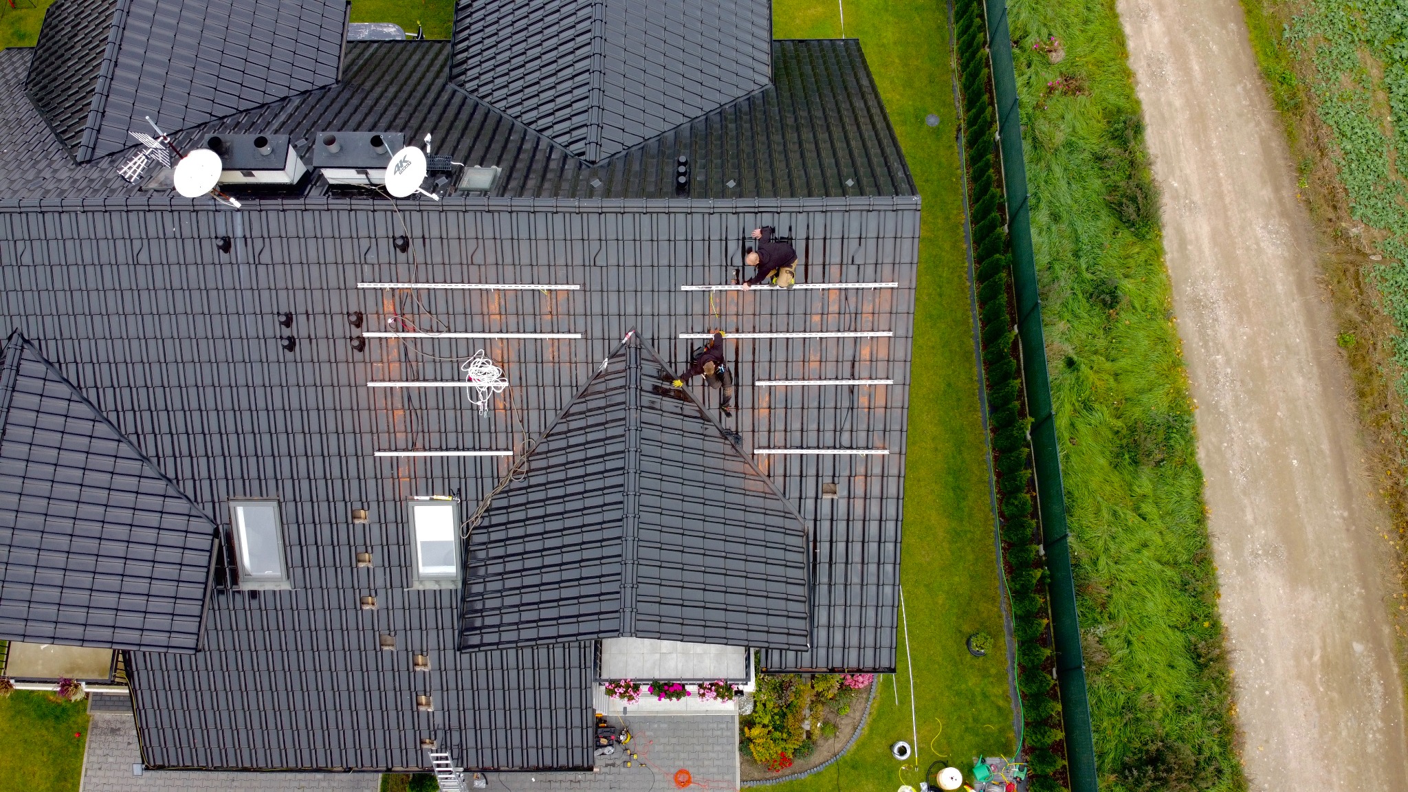 Widok z góry na montaż systemu szyn pod panele słoneczne na dachu domu pokrytego czarną dachówką, z widocznymi dwoma pracownikami w trakcie pracy, narzędziami i elementami instalacji.