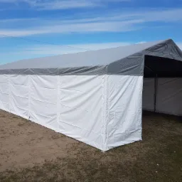 Rozłożony, biało-szary namiot plenerowy z wejściem, na trawiastym podłożu, z widocznym fragmentem białego płotu i czerwonego dachu budynku w tle.