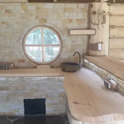 Wnętrze letniej kuchni z kamienia i drewna z okrągłym oknem, blatem z surowego drewna i kamiennym zlewem.