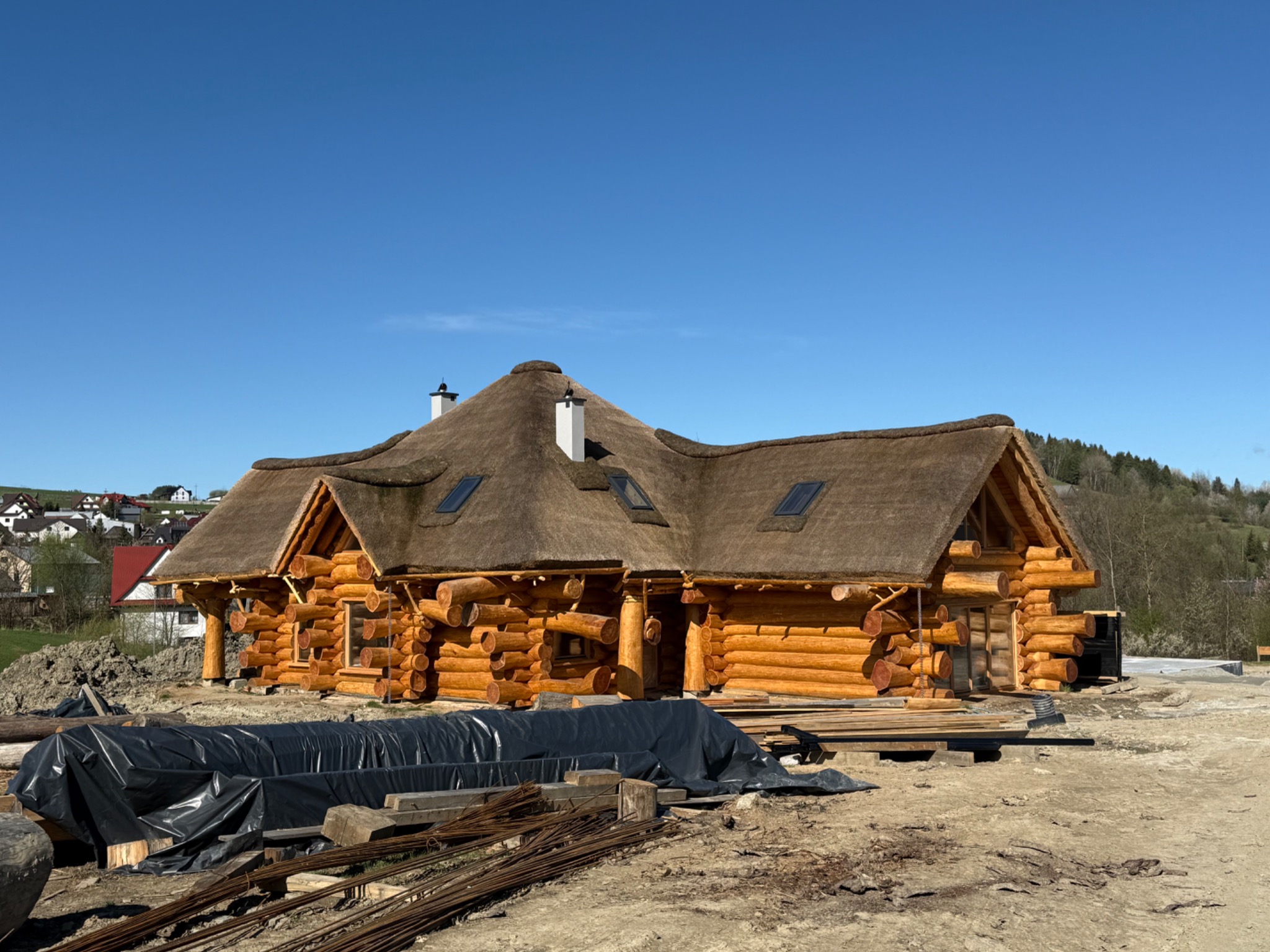 Wykończony dom z okrąglaków z trzcinowym dachem i dwoma kominami, widok z niskiej perspektywy na placu budowy z folią i prętami zbrojeniowymi.