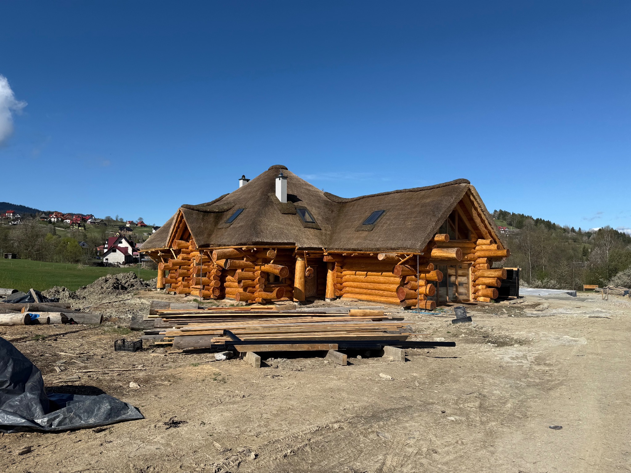 Surowy dom z bali z trzcinowym dachem i lukarnami w trakcie budowy, z widocznymi belkami i deskami na placu budowy, na tle błękitnego nieba i pagórkowatego krajobrazu.