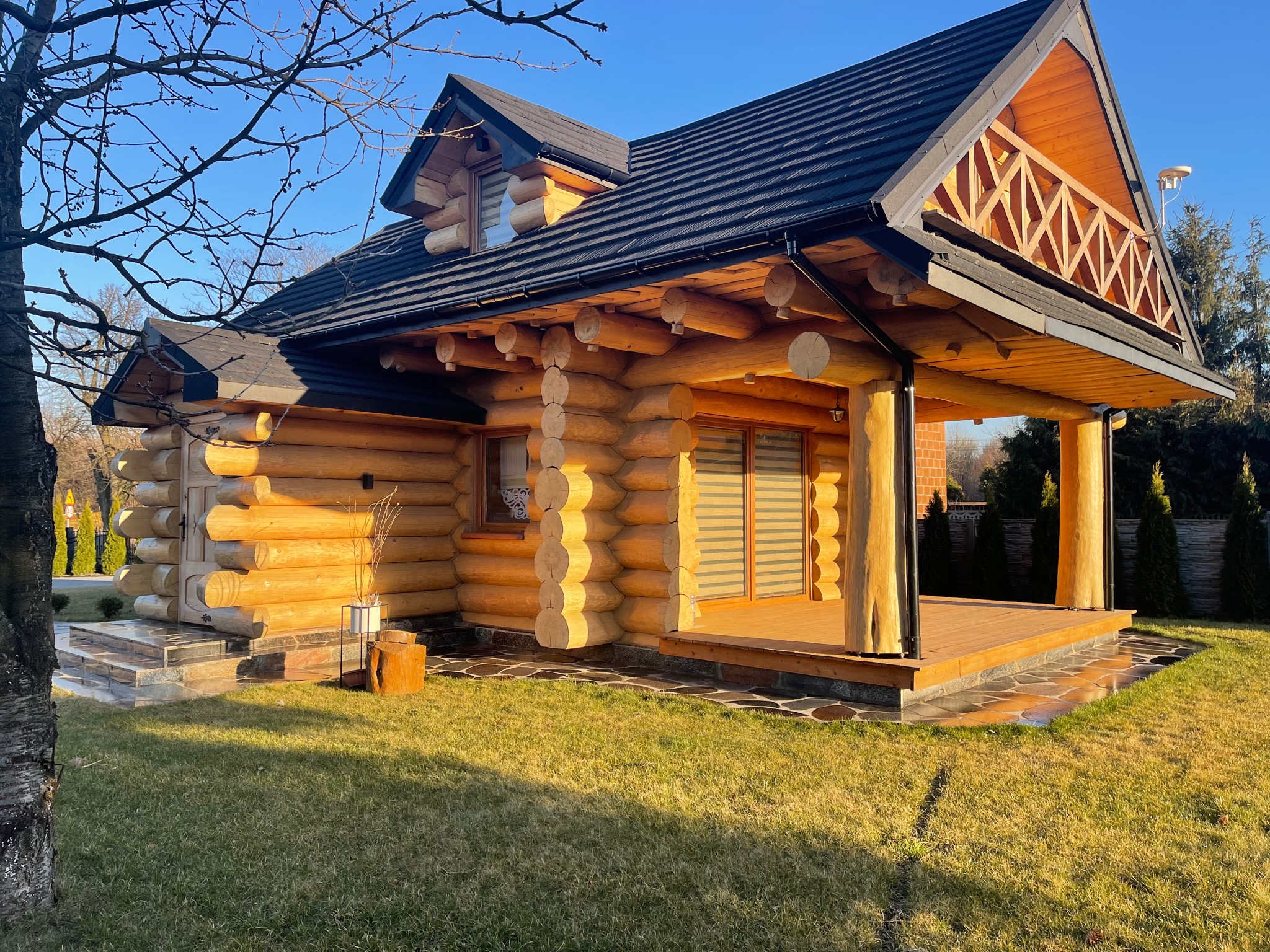 Nowoczesny dom z okrąglaków, z czarnym dachem i drewnianą werandą, otoczony zielonym trawnikiem w słoneczny dzień.