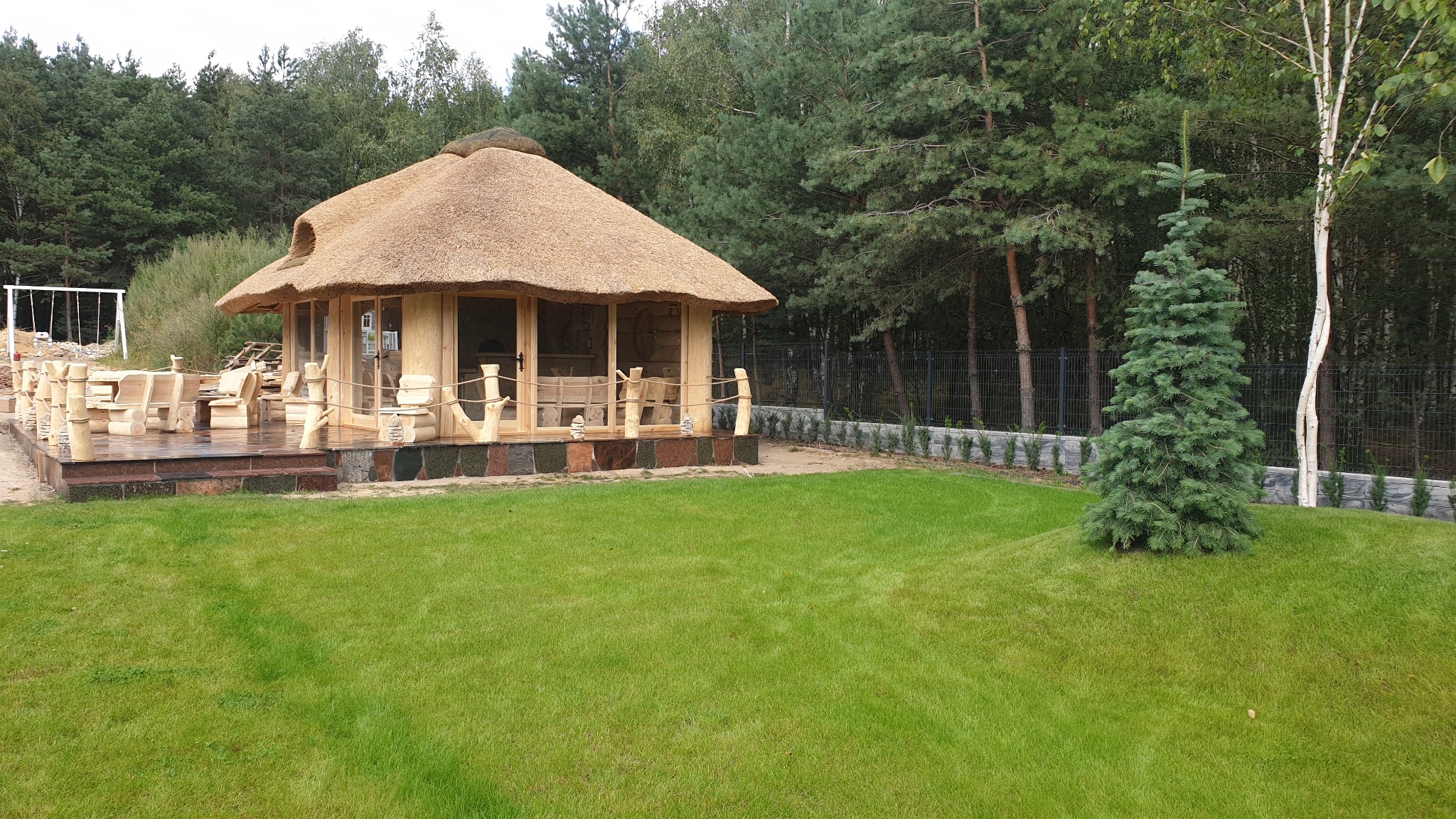 Ogród z drewnianą altaną krytą strzechą, wyposażoną w drewniane meble, otoczoną trawnikiem i drzewami.