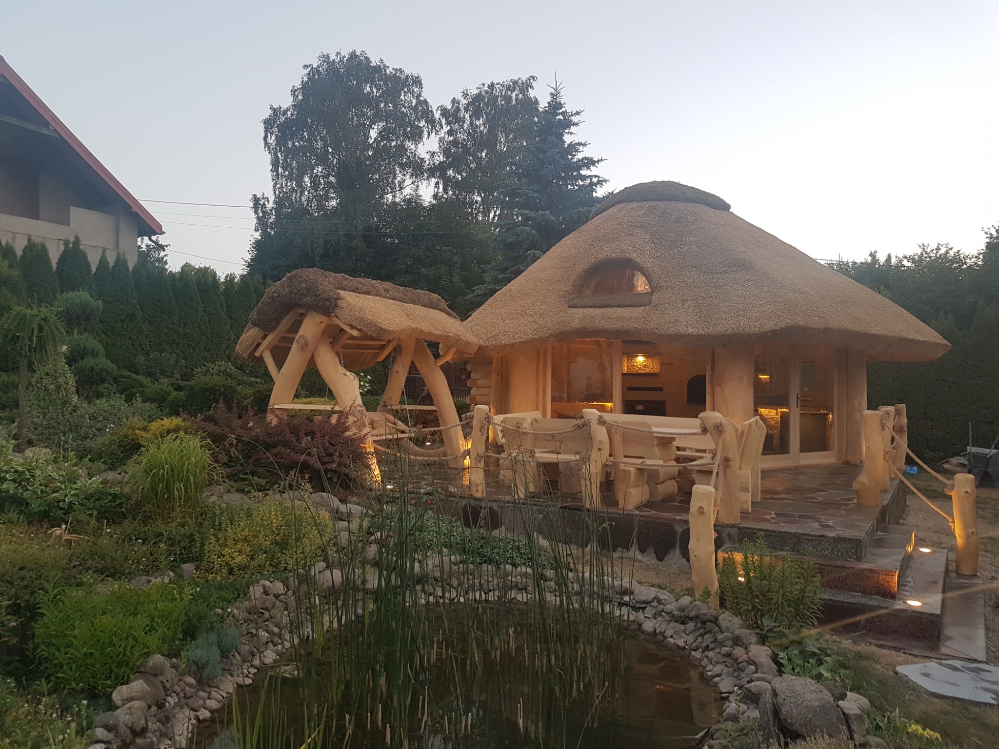 Altana ogrodowa z drewna z krytym strzechą dachem i huśtawką, otoczona oczkiem wodnym i roślinnością, oświetlona punktowymi lampami.