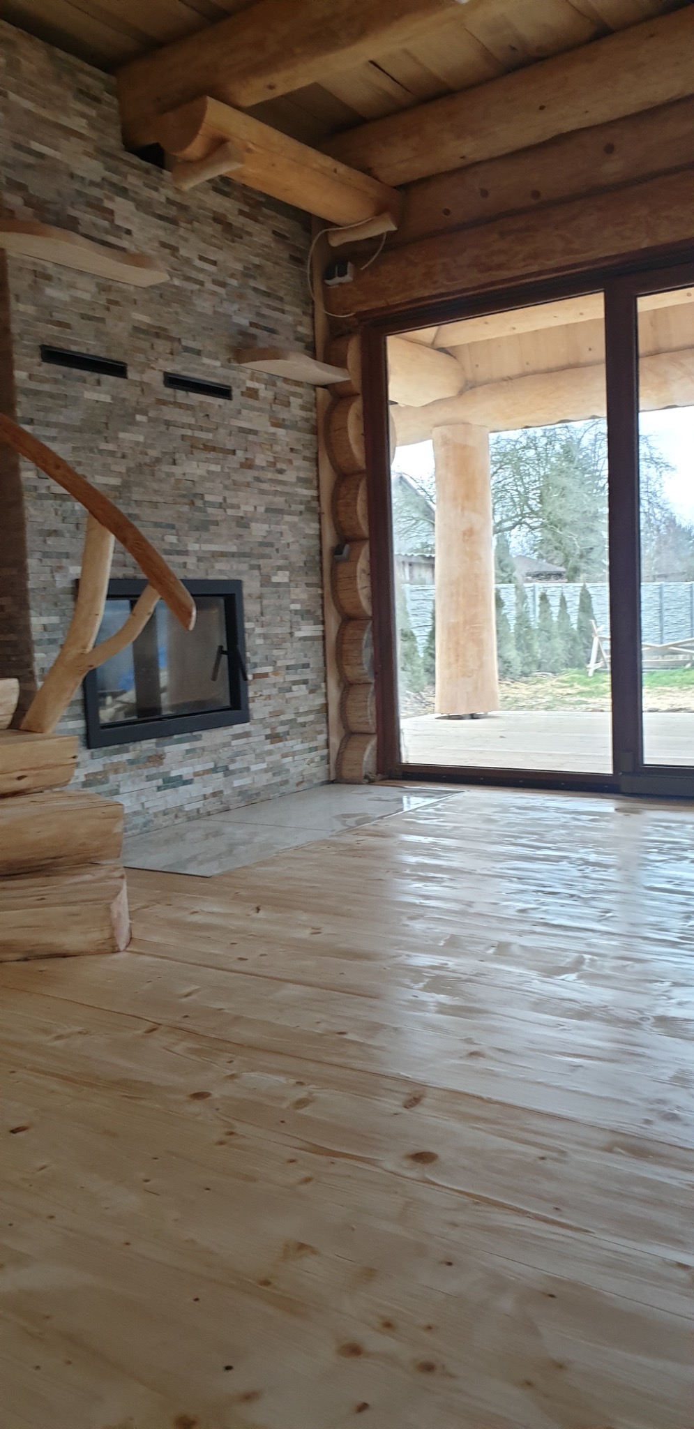 Wnętrze domu z drewnianymi belkami stropowymi, kamienną ścianą z kominkiem i drewnianymi schodami, widok na zewnątrz przez duże okno.
