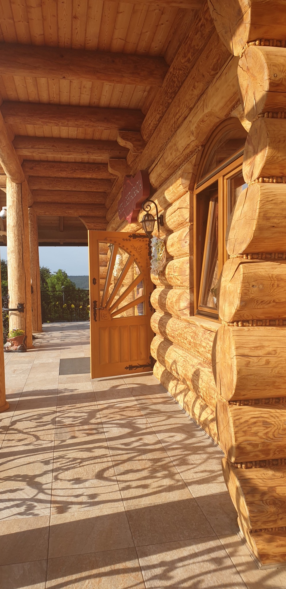 Zewnętrzny widok werandy domu z bali z otwartymi zdobionymi drzwiami i oknem, oświetlonej popołudniowym słońcem rzucającym cienie drzew na kafelkową podłogę.