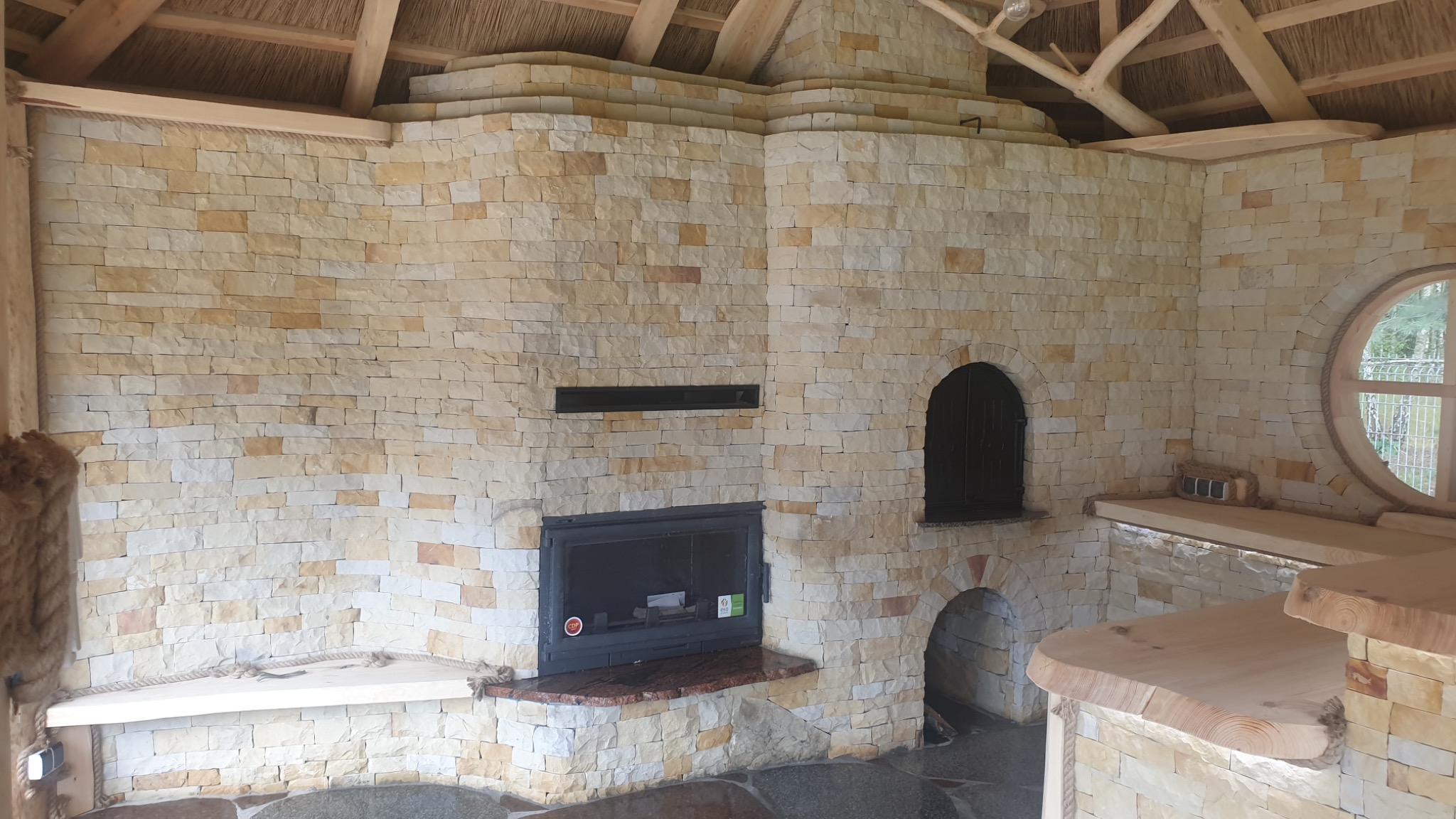 Wnętrze altany z kamienia z kominkiem, okrągłym oknem i drewnianą konstrukcją dachu krytego strzechą.