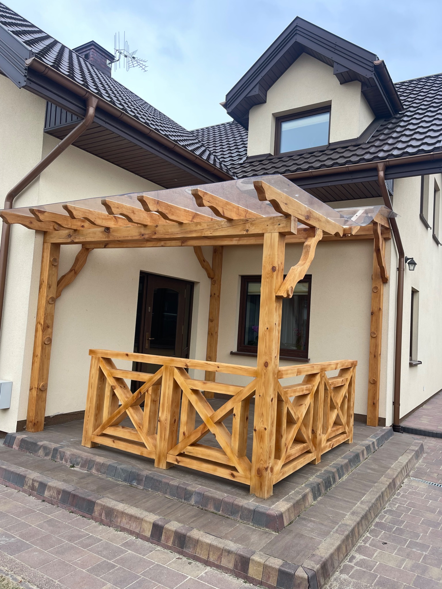 Drewniana pergola z przezroczystym zadaszeniem i ozdobną balustradą przed wejściem do domu z brązową stolarką okienną i dachówką.
