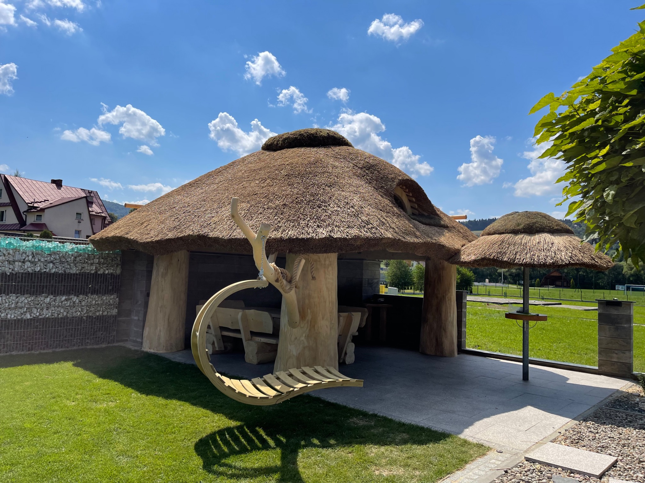 Oryginalna altana z krytym strzechą dachem i wiszącą huśtawką z drewna, otoczona zielonym trawnikiem w słoneczny dzień.