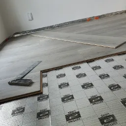 Montaż paneli podłogowych: widok z góry na podkład z nadrukowanymi liniami cięcia, częściowo ułożone panele o wzorze drewna i kątownik stolarski.