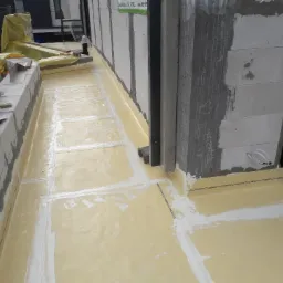 Żółta izolacja przeciwwodna na fundamencie budynku w trakcie prac budowlanych; widoczne bloczki betonowe i narzędzia murarskie.