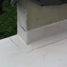 Fragment pokrycia dachowego z obróbką komina, widoczne ślady napraw i uszczelniania połączeń, w tle zieleń drzew.