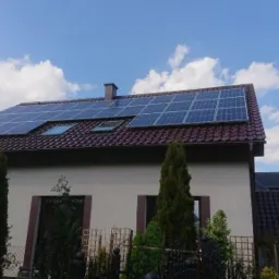 Dom jednorodzinny z brązową dachówką i zamontowanymi panelami fotowoltaicznymi oraz oknami dachowymi, na tle błękitnego nieba z chmurami.