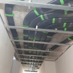 Montaż przewodów wentylacyjnych z czarnych rur karbowanych, mocowanych zielonymi uchwytami do sufitu podwieszanego z metalowej konstrukcji.