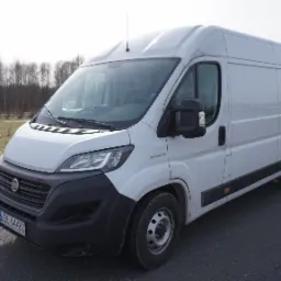 Biały samochód dostawczy Fiat Ducato zaparkowany na parkingu z widoczną tablicą rejestracyjną LBL 64480, drzewami w tle i słupami oświetleniowymi.