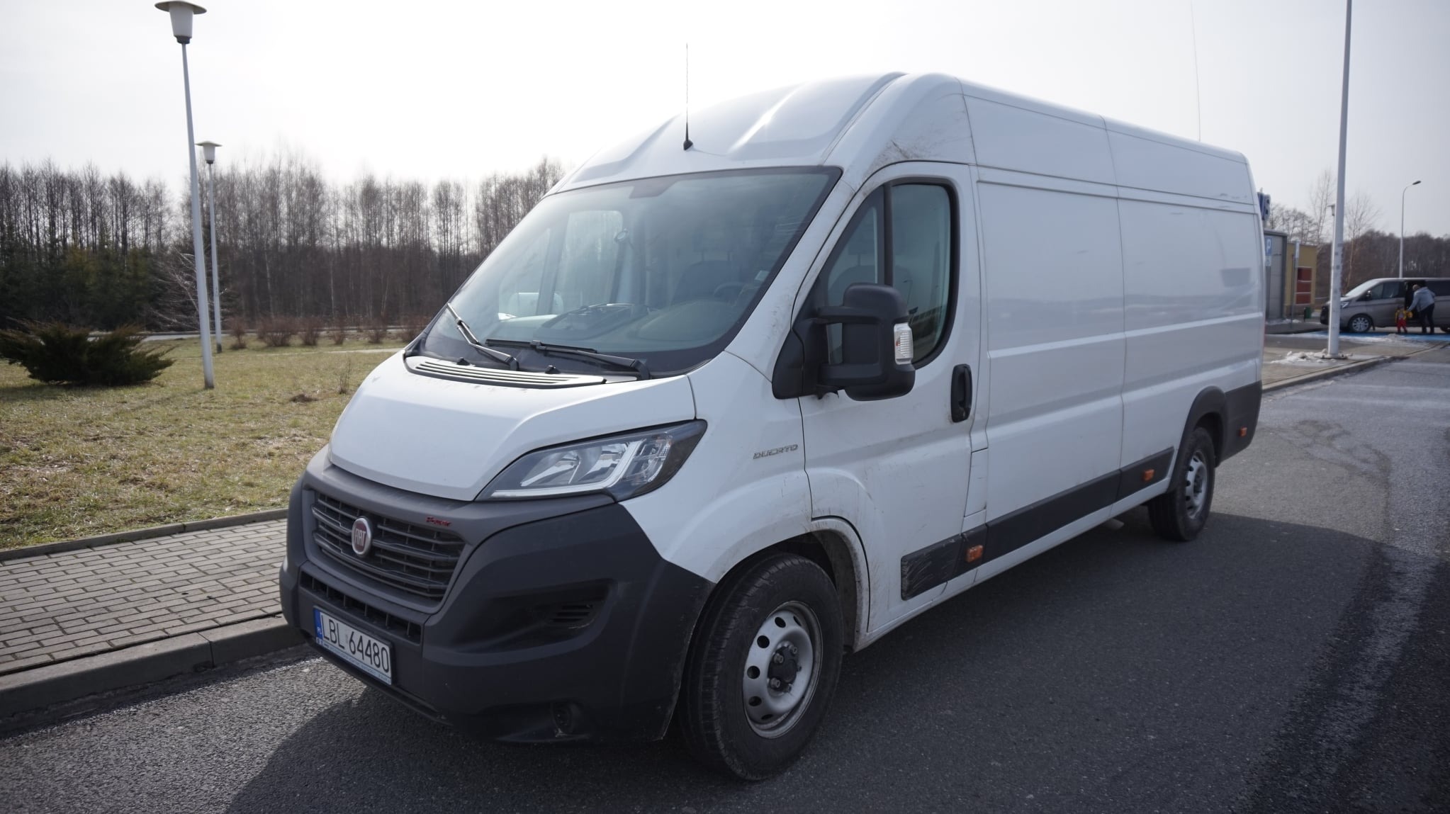 Biały samochód dostawczy Fiat Ducato zaparkowany na parkingu z widoczną tablicą rejestracyjną LBL 64480, drzewami w tle i słupami oświetleniowymi.