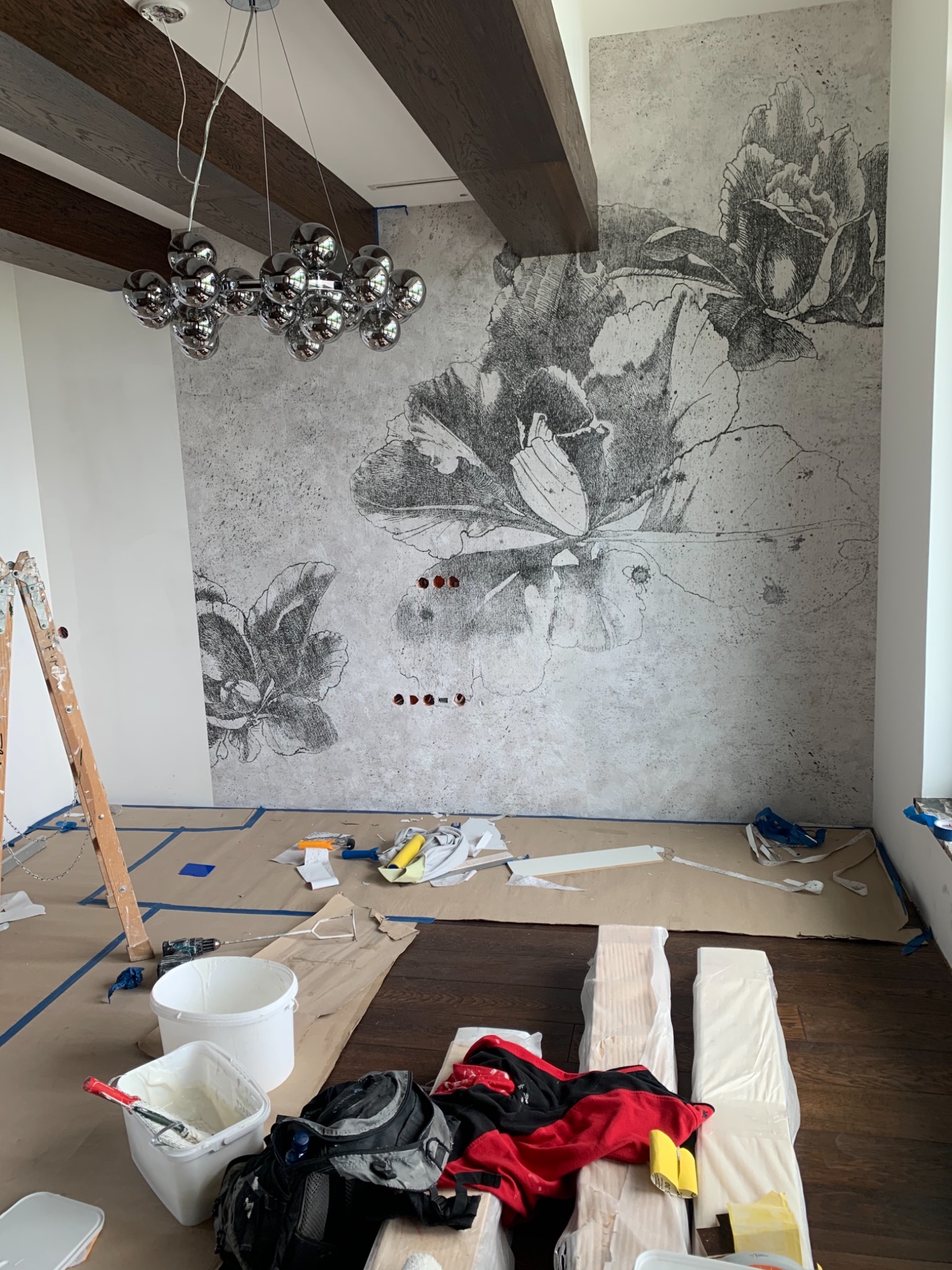 Ściana w trakcie dekorowania z motywem kwiatowym, widoczne narzędzia malarskie, drabina i zabezpieczenia podłogi, nowoczesna lampa wisząca w tle.