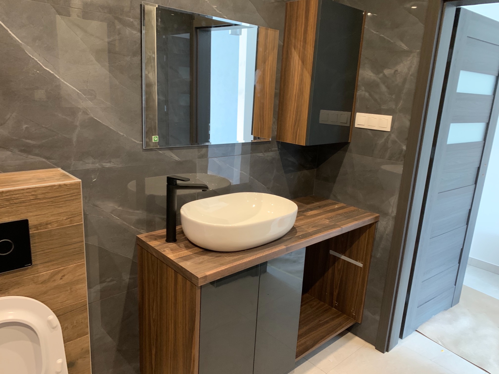 Nowoczesna łazienka z szarą marmurową płytką na ścianach, umywalką nablatową na szafce z drewnianym blatem, lustrem i drewnianą szafką wiszącą obok, oraz szarymi drzwiami.