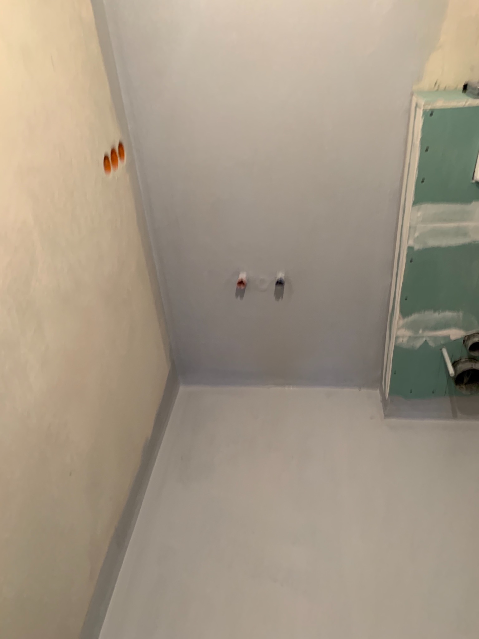 Świeżo pomalowane na szaro ściany i sufit w pomieszczeniu, widoczne otwory na instalacje elektryczne i fragment zabudowy z płyty gipsowo-kartonowej z widocznymi rurami.