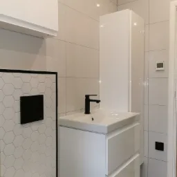 Nowoczesna łazienka z białymi płytkami, heksagonalnymi akcentami i czarną armaturą, minimalistyczna szafka pod umywalką z dwiema szufladami, wisząca szafka i zabudowa podtynkową toalety.