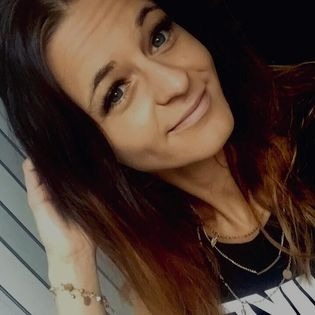 Portret uśmiechniętej brunetki z delikatnym makijażem, długimi rzęsami i subtelną biżuterią, pozującej na tle częściowo widocznych żaluzji.