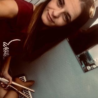 Uśmiechnięta brunetka z długimi włosami, trzymająca w dłoniach lokówkę, siedząca na tle niebieskiej ściany i ciemnej szafki.