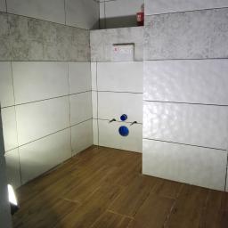 Marcin Modzelewski - Wykończona łazienka z białymi płytkami ściennymi o różnych wzorach i drewnopodobną podłogą, widoczne przyłącza wodne do toalety podtynkowej.