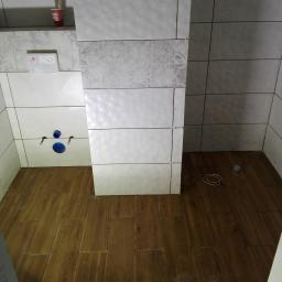 Marcin Modzelewski - Łazienka w trakcie wykańczania: częściowo ułożone płytki ścienne w odcieniach bieli i szarości z dekoracyjnym wzorem, drewnopodobne płytki podłogowe, widoczne elementy instalacji sanitarnej...