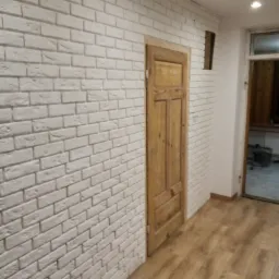 Ściana wyłożona białą cegłą dekoracyjną w pomieszczeniu z drewnianą podłogą i drzwiami, widoczna nieduża wnęka w ścianie.