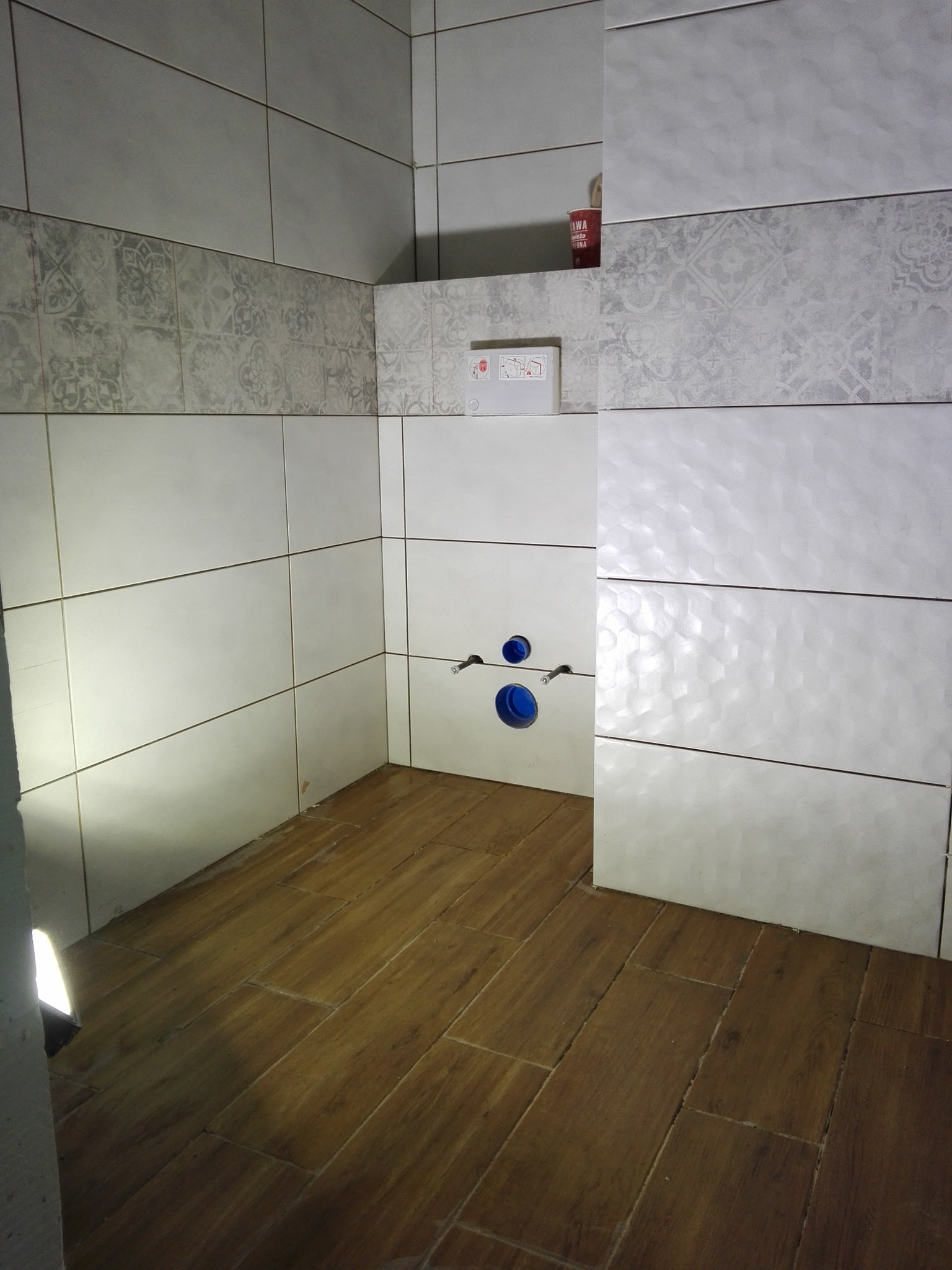 Wykończona łazienka z białymi płytkami ściennymi o różnych wzorach i drewnopodobną podłogą, widoczne przyłącza wodne do toalety podtynkowej.