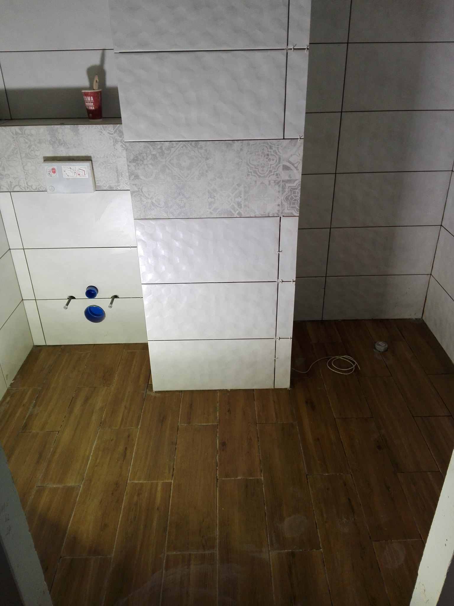 Łazienka w trakcie wykańczania: częściowo ułożone płytki ścienne w odcieniach bieli i szarości z dekoracyjnym wzorem, drewnopodobne płytki podłogowe, widoczne elementy instalacji sanitarnej...