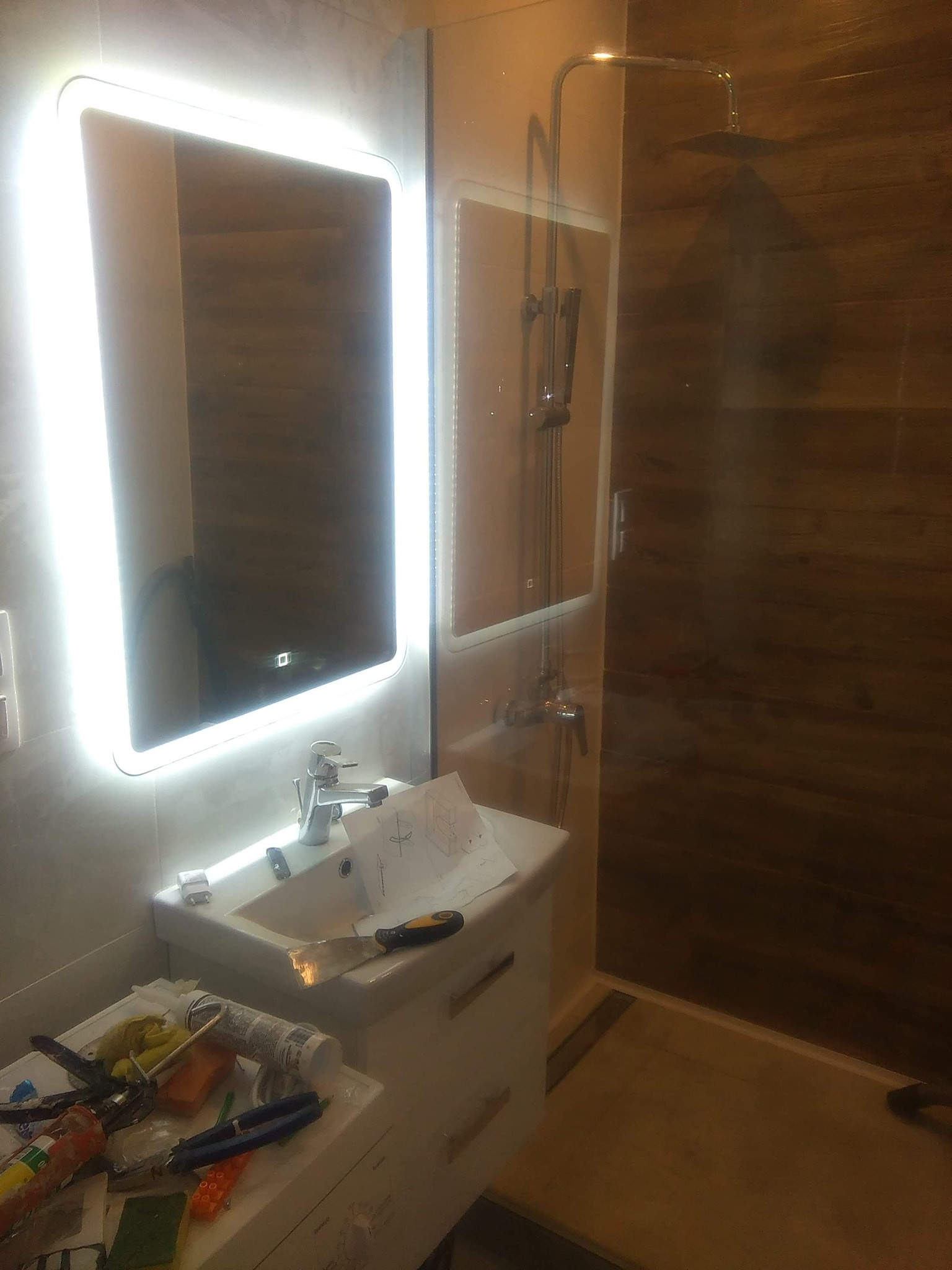 Nowoczesna łazienka w trakcie wykańczania: podświetlane lustro LED, kabina prysznicowa z deszczownicą i narzędzia glazurnicze na blacie umywalki.