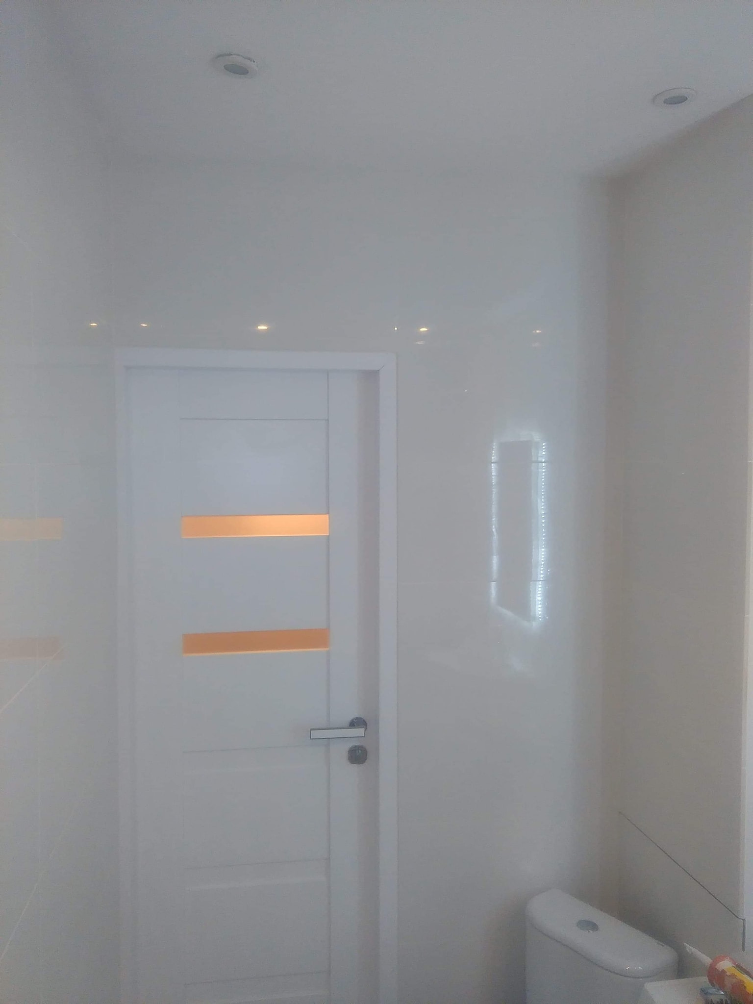 Biała łazienka z błyszczącymi płytkami na ścianach, białą drzwiami z dwoma poziomymi wstawkami w kolorze miedzianym, podwieszanym sufitem z oświetleniem punktowym oraz białą toaletą w rogu.