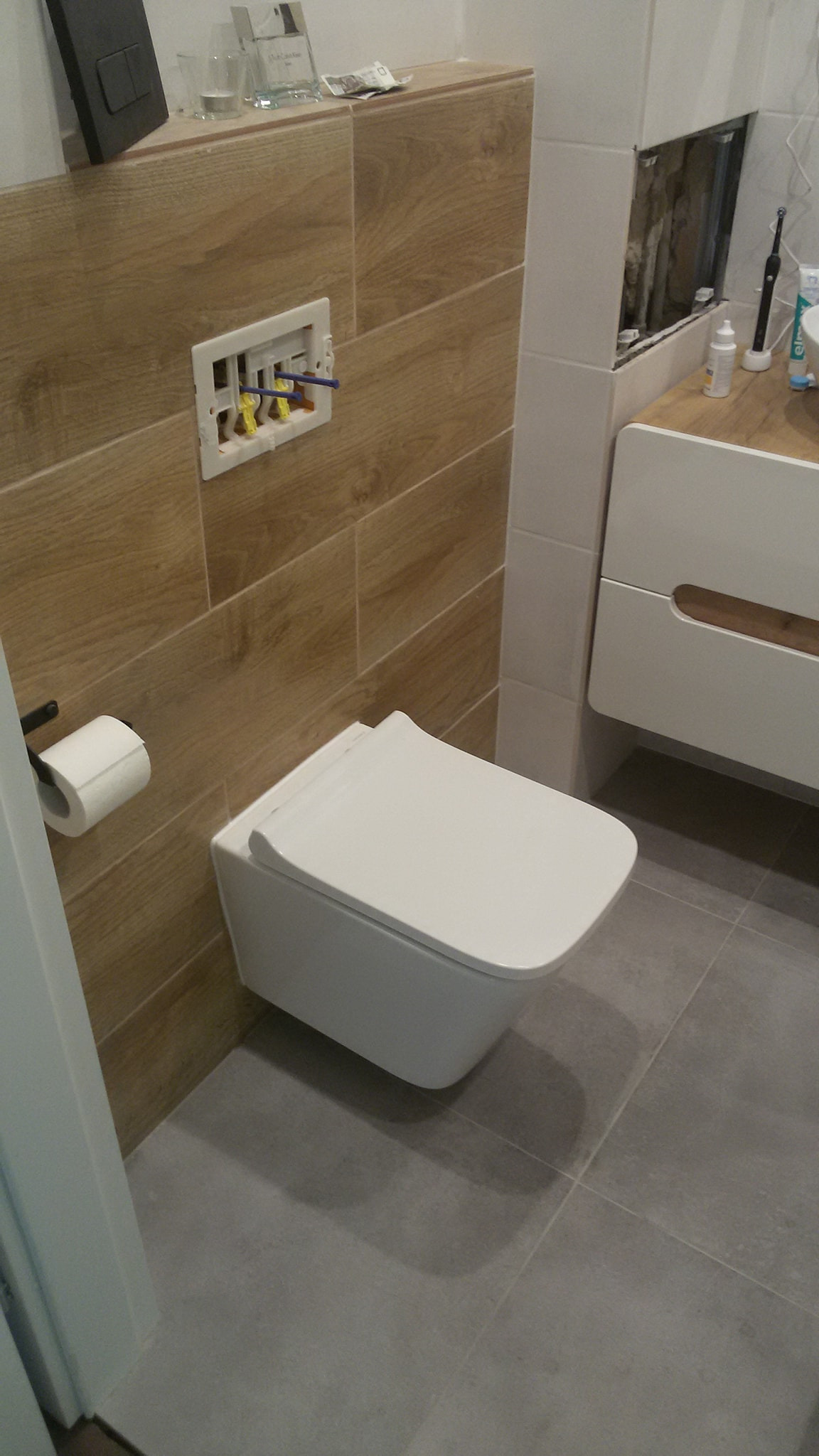 Wisząca miska WC w łazience z drewnopodobnymi płytkami na ścianie, szarymi płytkami na podłodze i częściowo wykończonym otworem w ścianie, sugerującym trwający remont.