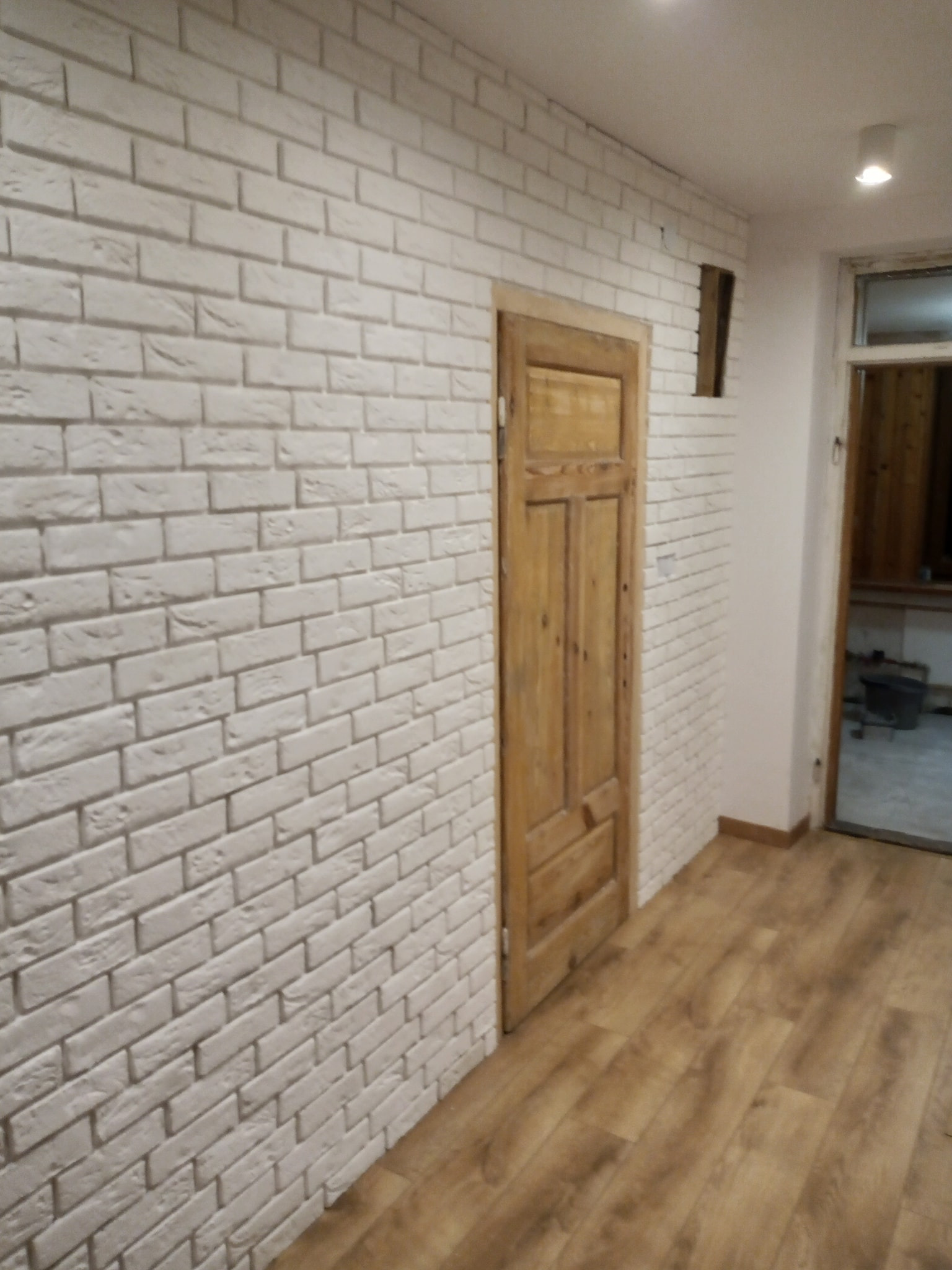 Ściana wyłożona białą cegłą dekoracyjną w pomieszczeniu z drewnianą podłogą i drzwiami, widoczna nieduża wnęka w ścianie.