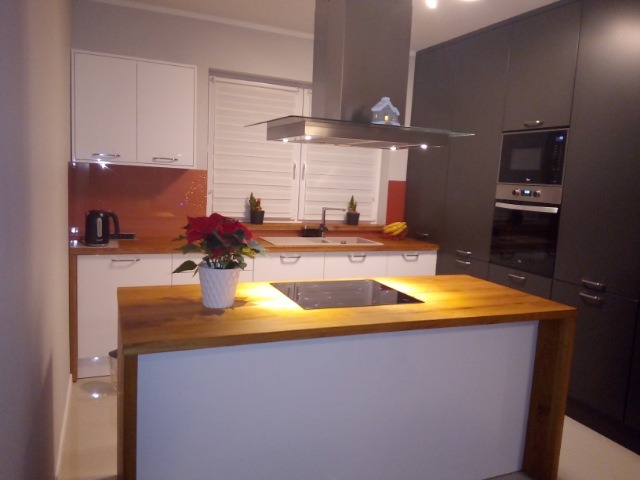 Nowoczesna kuchnia z wyspą o drewnianym blacie, na której stoi doniczka z poinsecją, w tle białe i grafitowe szafki oraz okap nad płytą grzewczą.