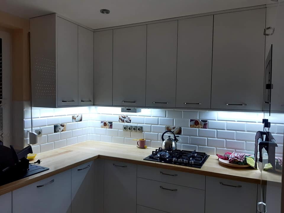 Jasnoszara zabudowa kuchenna z drewnianym blatem, oświetleniem LED pod szafkami i płytkami w stylu metro z dekoracyjnymi akcentami.