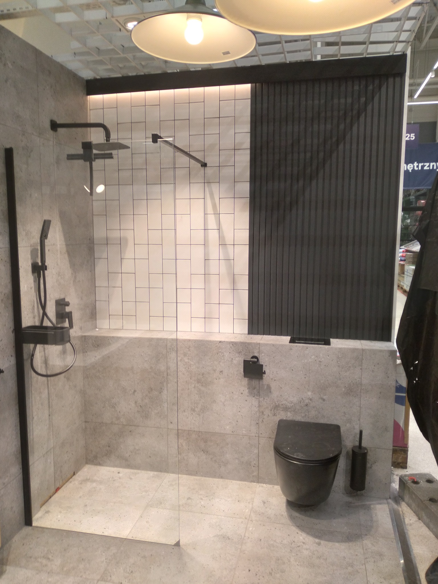 Nowoczesna łazienka z szarą armaturą, kabiną prysznicową z białymi płytkami i czarnym panelem dekoracyjnym, podwieszana toaleta w kolorze grafitowym.