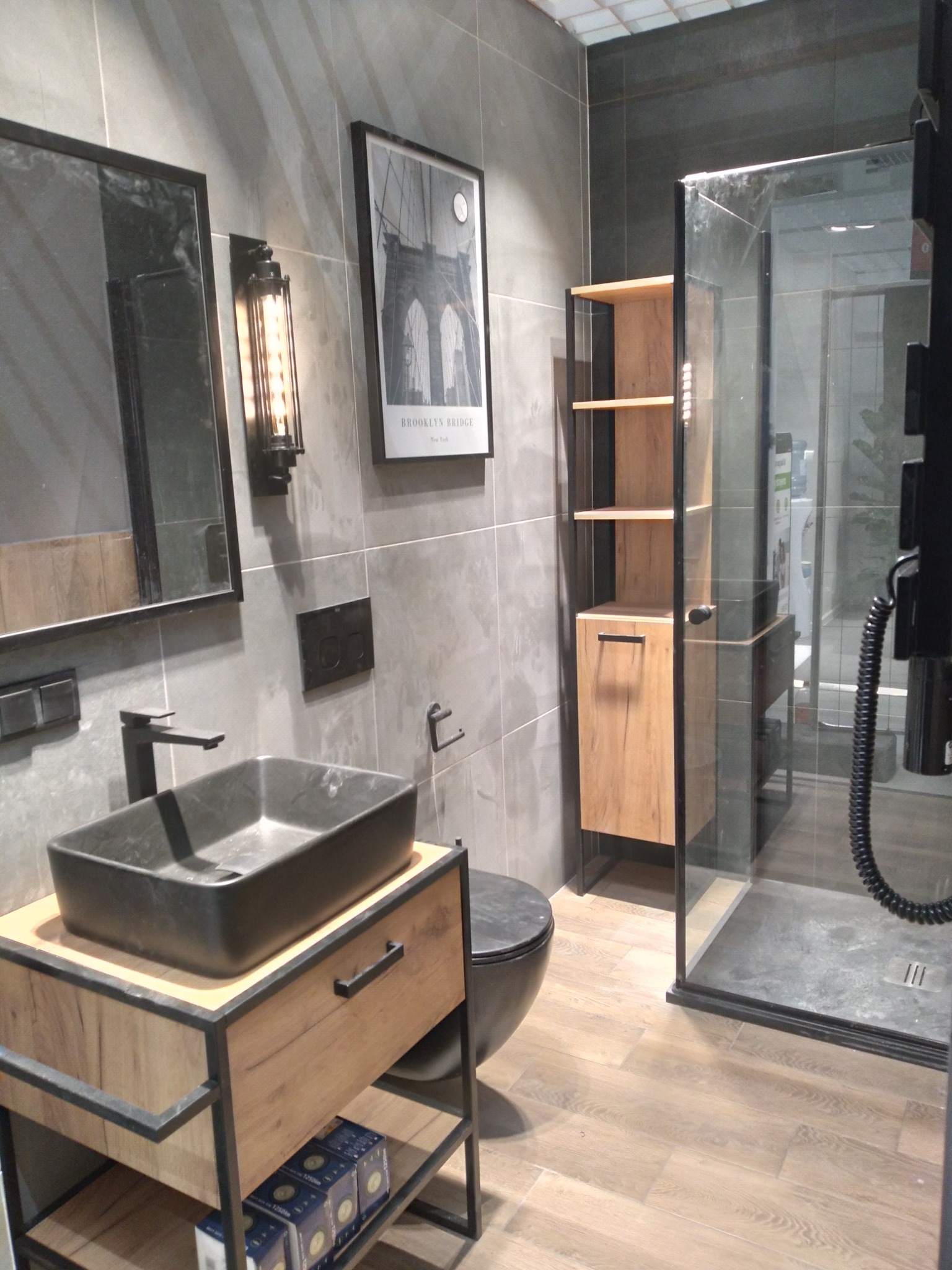 Nowoczesna łazienka z szarą glazurą, czarną umywalką nablatową na szafce z drewnopodobnymi frontami, minimalistyczną lampą ścienną i kabiną prysznicową typu walk-in.