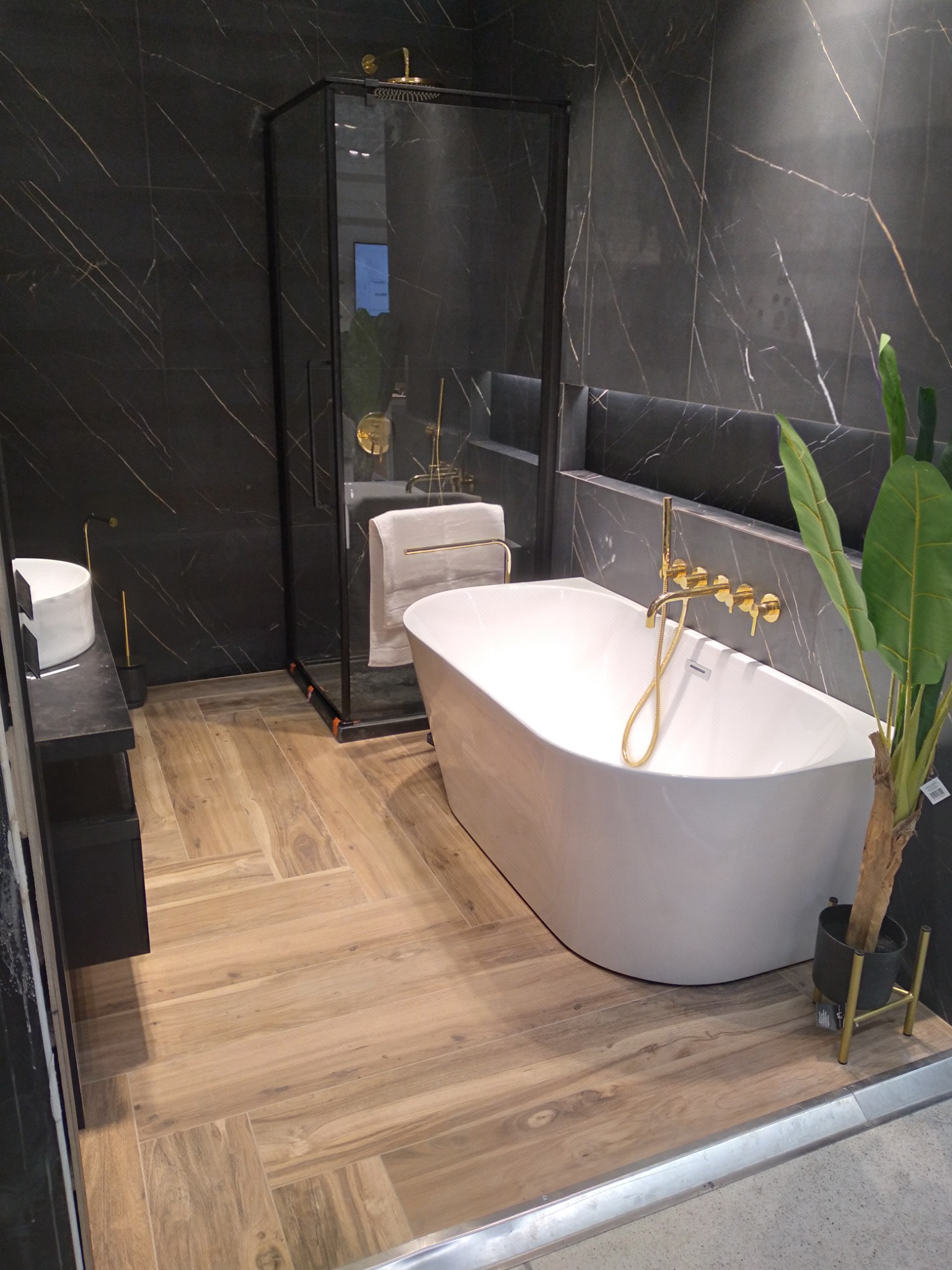 Nowoczesna łazienka z czarnymi płytkami imitującymi marmur, wolnostojącą białą wanną z złotą armaturą, kabiną prysznicową ze złotymi detalami i drewnopodobną podłogą ułożoną w jodełkę.