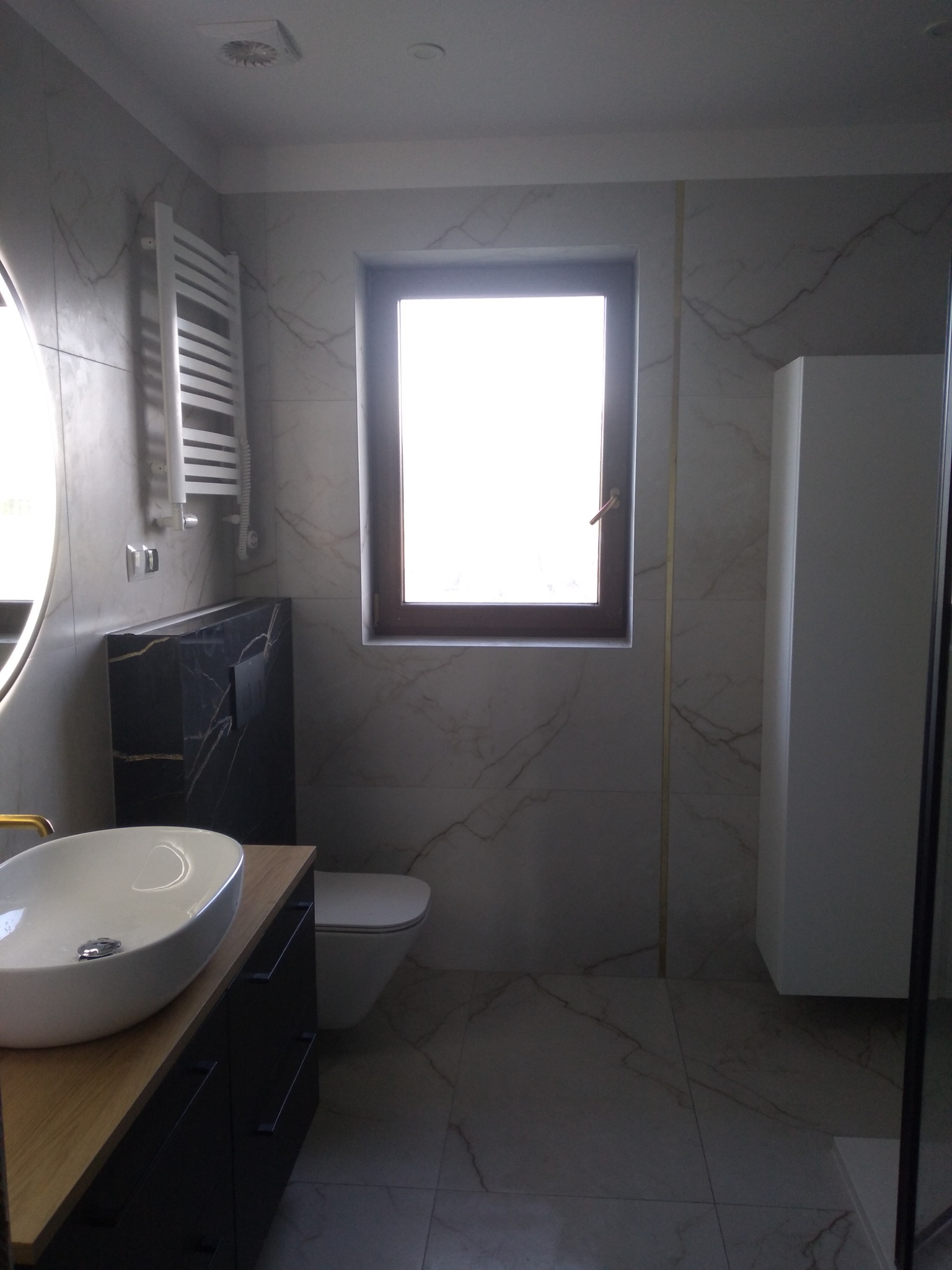 Nowoczesna łazienka z marmurowymi płytkami, umywalką nablatową na drewnianym blacie, podwieszaną toaletą i grzejnikiem drabinkowym.