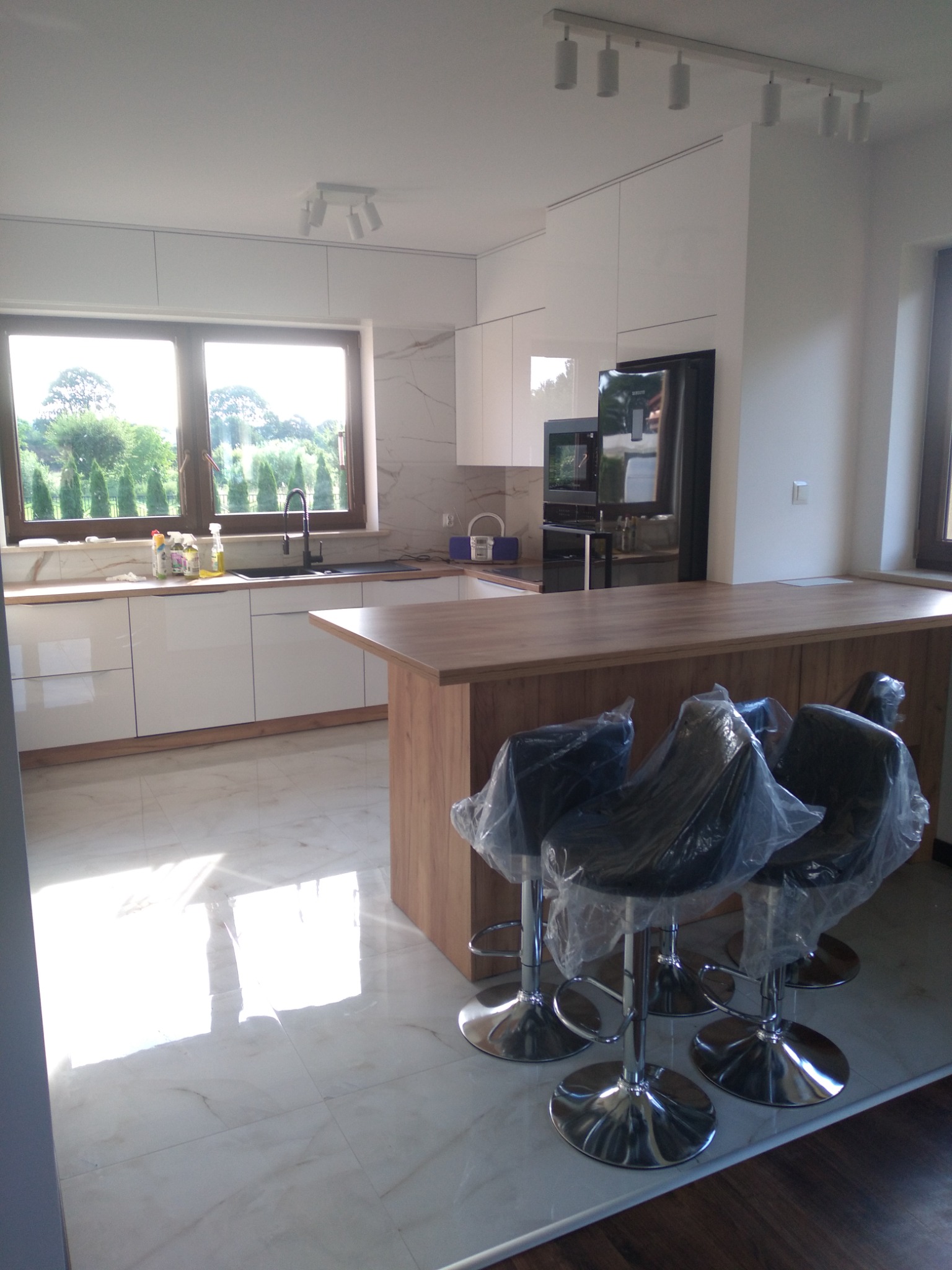 Nowoczesna kuchnia z białymi szafkami, marmurową podłogą i drewnianym blatem, trzy krzesła barowe owinięte folią ochronną, widok na zieleń za oknem.