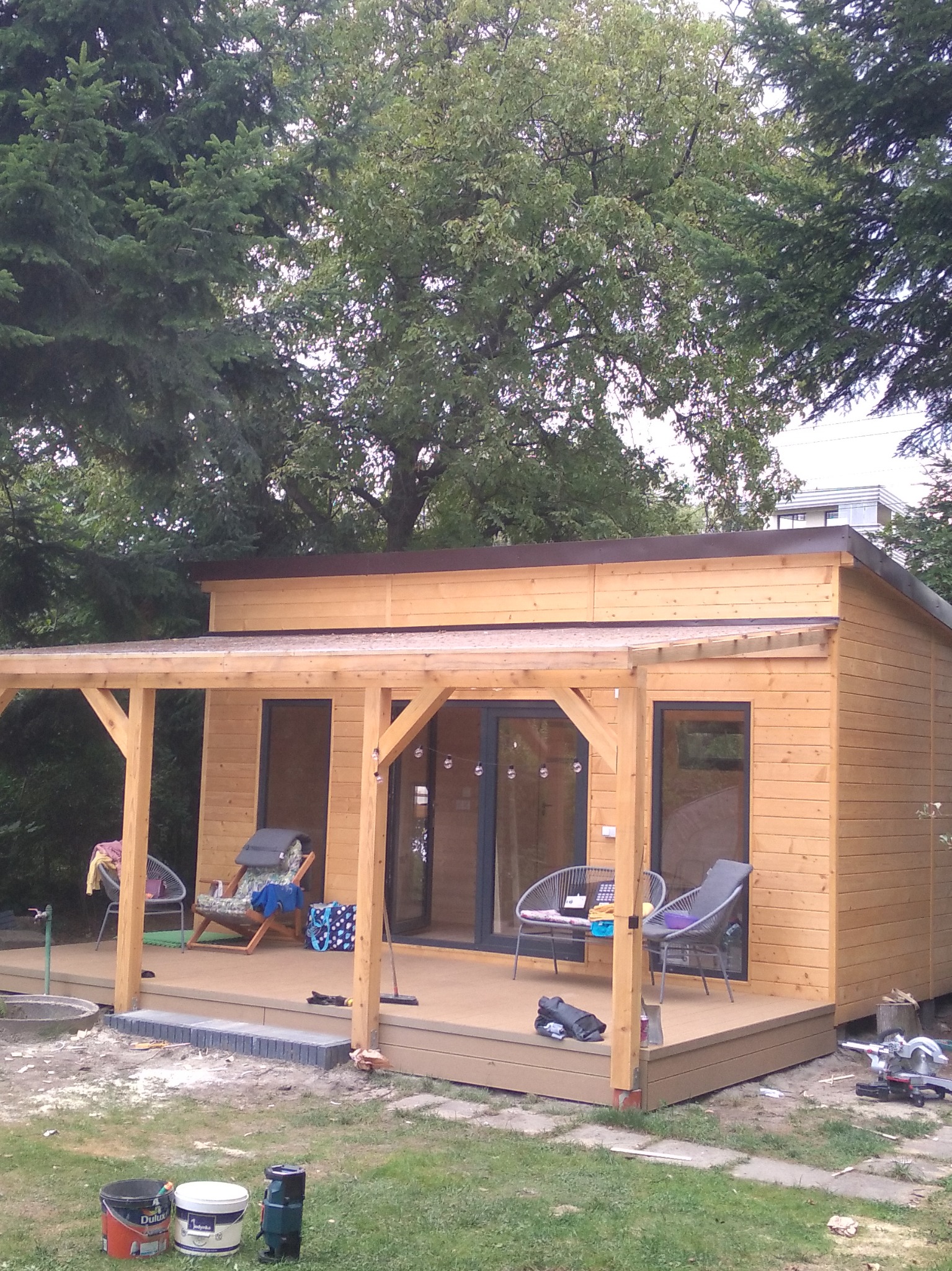 Nowoczesny drewniany domek letniskowy z zadaszonym tarasem, otoczony zielenią, z widocznymi narzędziami i akcesoriami na tarasie, sugerującymi trwające prace wykończeniowe.