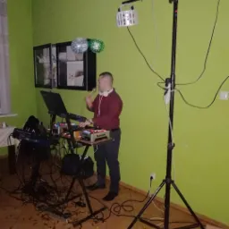 DJ w trakcie występu, obsługujący sprzęt Numark, z oświetleniem scenicznym na statywie, na podłodze rozsypane konfetti, w tle zielona ściana i szafka z dekoracjami.